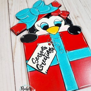 A red and blue penguin wearing a blue bow peeking out of a red gift box with a blue ribbon. The gift box has a white tag that says 'Seasons Greetings'.