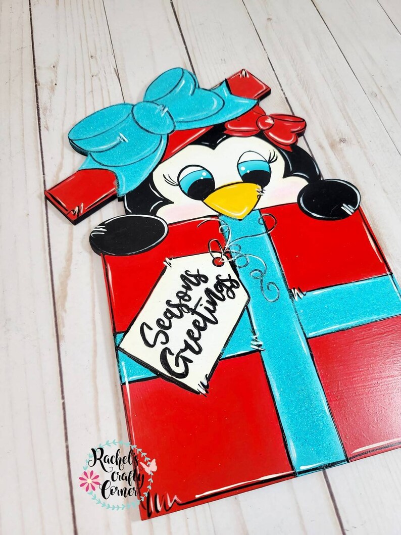A red and blue penguin wearing a blue bow peeking out of a red gift box with a blue ribbon. The gift box has a white tag that says 'Seasons Greetings'.
