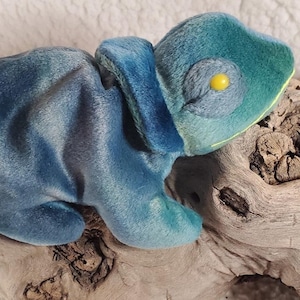 May include: A blue and green plush chameleon toy with yellow eyes. The chameleon is sitting on a piece of driftwood.