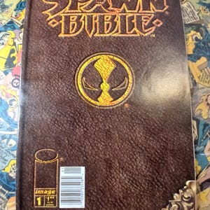 May include: A comic book titled "Spawn Bible" with a brown leather-like cover. The title is in gold lettering, and a gold emblem is centered on the cover. The comic book is issue number 1, with a price of $1.95.