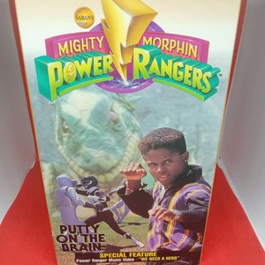 May include: A VHS tape cover for the Mighty Morphin Power Rangers episode "Putty on the Brain". The cover features a yellow lightning bolt with the words "Mighty Morphin Power Rangers" in green and purple letters. The cover also features a photo of the Black Ranger, Zack, in a purple and yellow shirt, performing a karate move. The text "Special Feature: Power Ranger Music Video - "We Need a Hero" is also on the cover.
