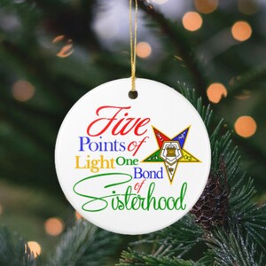 Puede incluir: Un adorno redondo de cerámica blanca con el texto Five Points of Light One Bond of Sisterhood en letras rojas, azules, amarillas y verdes. A la derecha, se muestra un emblema de estrella multicolor que contiene varios símbolos internos.