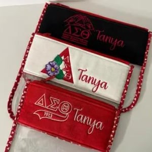 May include: Three zippered pouches with different colored fabric and embroidery. The pouches are red, white, and black. Each pouch has the letters "&Delta;&Sigma;&Theta;" embroidered on it, along with the name "Tanya".