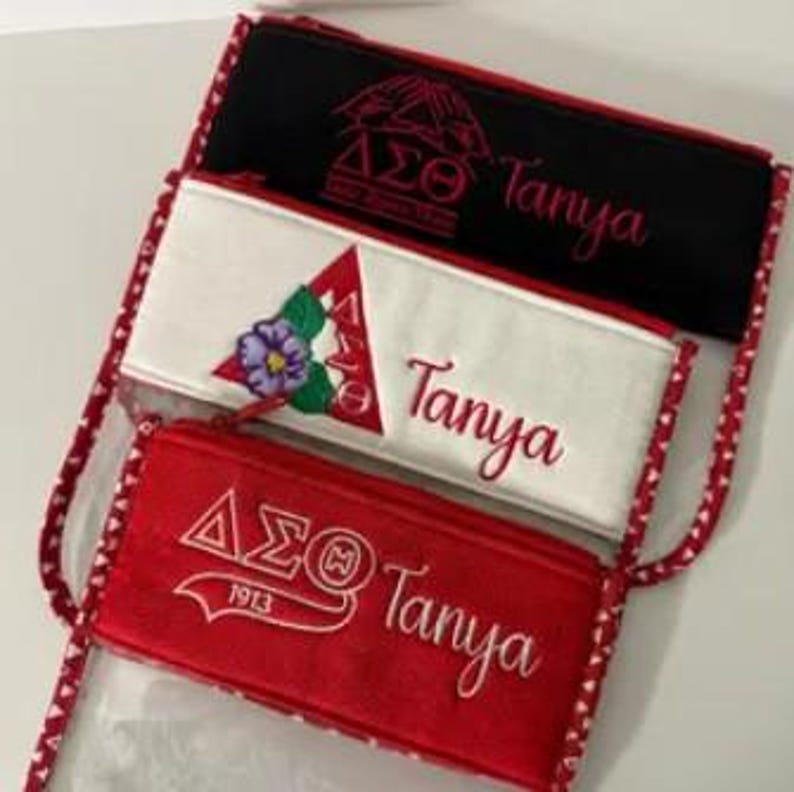 May include: Three zippered pouches with different colored fabric and embroidery. The pouches are red, white, and black. Each pouch has the letters "&Delta;&Sigma;&Theta;" embroidered on it, along with the name "Tanya".