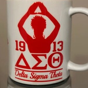 May include: White ceramic mug with a red silhouette of a person with arms raised in a triangle shape. The mug has the text "1913 &Delta;&Sigma;&Theta; Delta Sigma Theta" in red.