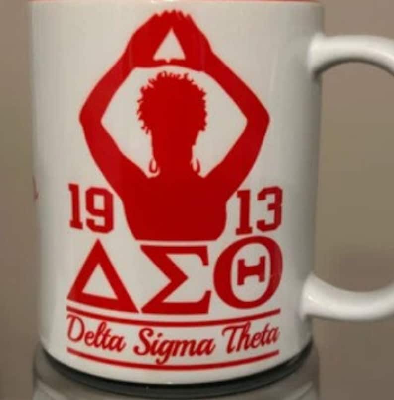 May include: White ceramic mug with a red silhouette of a person with arms raised in a triangle shape. The mug has the text "1913 &Delta;&Sigma;&Theta; Delta Sigma Theta" in red.