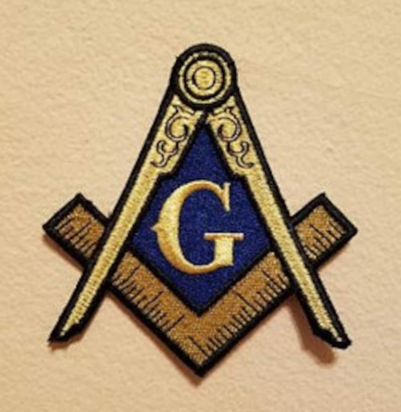 May include: Embroidered patch depicting the Masonic Square and Compasses symbol. The patch features a blue centre with a gold "G", framed by gold and black detailing. The background is a light tan colour.