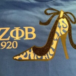 May include: Blue t-shirt featuring a high-heeled shoe design with a black glitter outline and a gold heel. The shoe has a blue and white pattern. The text "Z&Phi;B 1920" is printed in gold.