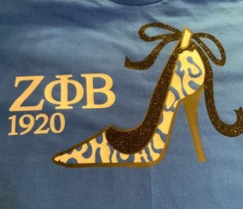 May include: Blue t-shirt featuring a high-heeled shoe design with a black glitter outline and a gold heel. The shoe has a blue and white pattern. The text "Z&Phi;B 1920" is printed in gold.