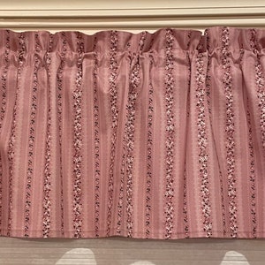 May include: A dusty rose valance with vertical stripes and floral accents. The fabric is gathered at the top, creating a ruffled effect. The curtain rod is white.