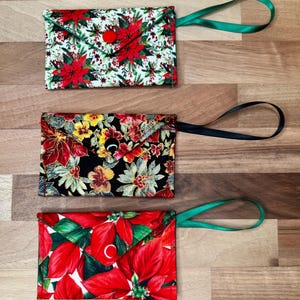May include: Three fabric pouches with floral patterns. The top pouch has a white background with red poinsettias and a green ribbon loop. The middle pouch has a black background with red and yellow flowers and a black ribbon loop. The bottom pouch has a red background with green leaves and a green ribbon loop.