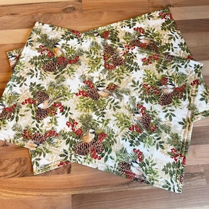 May include: Two rectangular placemats with a repeating pattern of birds, pinecones, and red berries on a cream background. The design includes green foliage and is set against a light cream backdrop. The placemats are made of a soft, possibly fabric material.