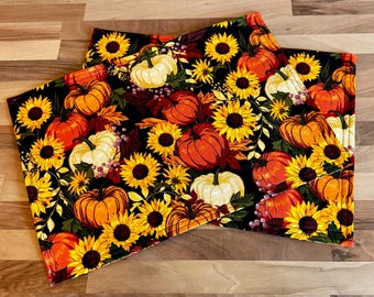 May include: Two rectangular placemats with a black background, featuring a vibrant autumn design. The pattern includes orange and white pumpkins, yellow sunflowers, and fall foliage. The placemats are likely made of fabric.