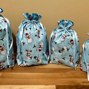 May include: Four light blue fabric gift bags with a snowman pattern. The bags are tied with light blue ribbon and have a ruffled top. The snowman design includes snowmen with red hats and scarves.