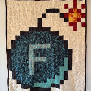May include: A handmade quilt featuring a pixelated bomb design with a teal body and a red and yellow explosion. The letter "F" is in the center of the bomb. The quilt has a brown border and is hanging on a white wall.