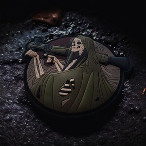 May include: A green and brown embroidered patch depicting a skeleton wearing a hooded cloak and holding a rifle. The patch has a detailed design and is perfect for adding a unique touch to clothing or bags.