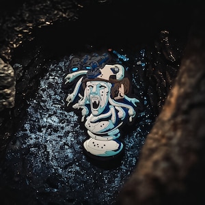 May include: A blue and white enamel pin depicting a Medusa-like figure with snakes for hair, wearing a helmet with a night vision scope. The figure is screaming with its mouth open and fangs visible.