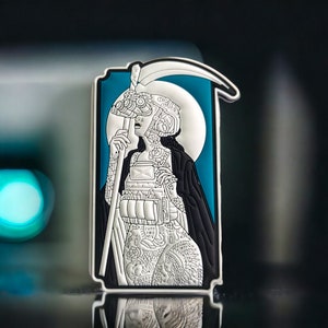 May include: A silver and black enamel pin featuring a stylized image of a woman holding a scythe. The woman is wearing a helmet and has a detailed tattoo design on her body. The background is a teal blue color.