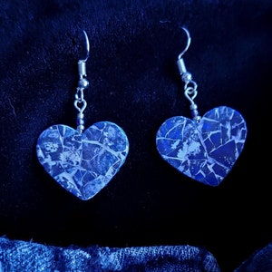 Puede incluir: Un par de pendientes en forma de corazón con un patrón de mármol azul y blanco. Los pendientes están hechos de metal y tienen un acabado plateado. Los pendientes cuelgan de un fondo de mezclilla azul con una etiqueta que dice "hecho a mano con amor".
