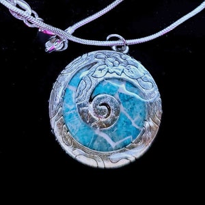 May include: A silver necklace with a round pendant featuring a blue and white swirl design. The pendant has a detailed silver border with a floral pattern.