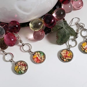 May include: Three pairs of silver earrings with colorful mosaic-like round pendants. The earrings have a simple design with a small hoop and a dangling pendant.