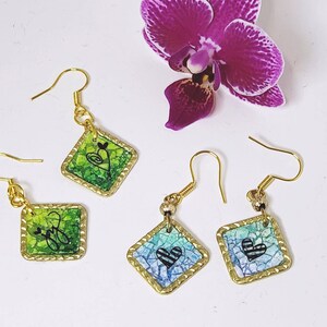 May include: Three pairs of gold earrings with a diamond-shaped pendant. Each pendant features a different coloured mosaic design with a black heart in the centre.