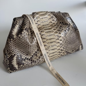 May include: A clutch bag with a snakeskin pattern in shades of grey, brown, and cream. The bag has a gathered top and a cream-coloured strap with the text "HEDONISTBALI" printed on it. The bag is made of a textured material.