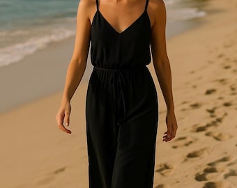 May include: A woman wearing a black sleeveless jumpsuit with a tie at the waist, walking on a sandy beach. She is wearing black sunglasses.