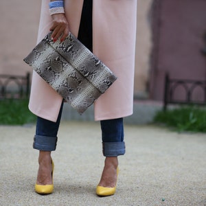 May include: A woman is wearing a pink coat, blue jeans, and yellow heels. She is holding a snakeskin clutch.