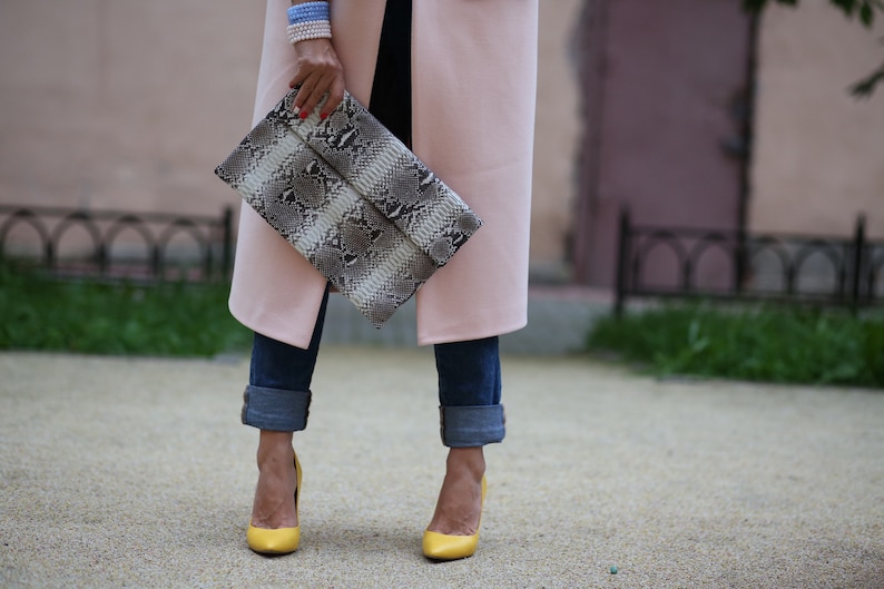 May include: A woman is wearing a pink coat, blue jeans, and yellow heels. She is holding a snakeskin clutch.
