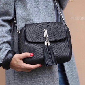 May include: Black leather crossbody bag with a flap closure, silver chain strap, and decorative tassels. The bag has a textured, snakeskin-like pattern. The text "#HEDONIST" is visible on the right side of the image.