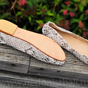 May include: A pair of ballet flats with a snakeskin pattern in shades of gray and white. The soles are light brown, and the size 38 is visible on the sole. The shoes are displayed on a weathered wooden surface.