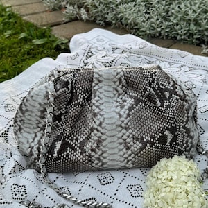 May include: A white and black snakeskin clutch bag with a silver chain strap. The bag is sitting on a white crocheted tablecloth.