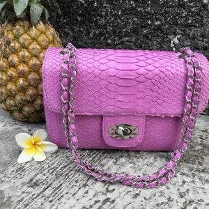 K&ouml;nnte beinhalten: Eine kleine, helllila Handtasche aus Schlangenleder mit einer silbernen Kettenkette und einem silbernen Verschluss.