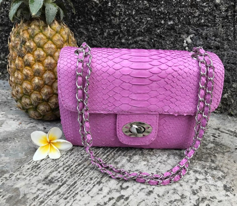 K&ouml;nnte beinhalten: Eine kleine, helllila Handtasche aus Schlangenleder mit einer silbernen Kettenkette und einem silbernen Verschluss.