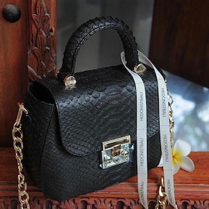 May include: A black leather handbag with a snakeskin pattern and a gold chain strap. The bag has a gold lock and a white ribbon with the text "HEDONISTBALI" printed on it.