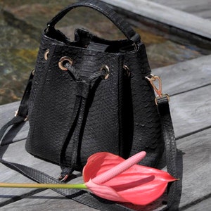 May include: A black faux leather bucket bag with a snakeskin pattern and a long strap. The bag has a drawstring closure and gold-toned hardware. A pink flower is in the foreground.