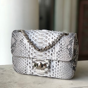 Może przedstawiać: Mała biała torebka ze skóry węża z srebrnym łańcuszkiem i srebrnym zapięciem.