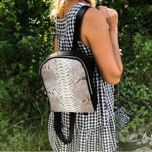 以下が含まれることがあります： 白いヘビの皮の模様がついた黒い革のバックパック。バックパックはジッパーで閉じて、調節可能なストラップが付いています。