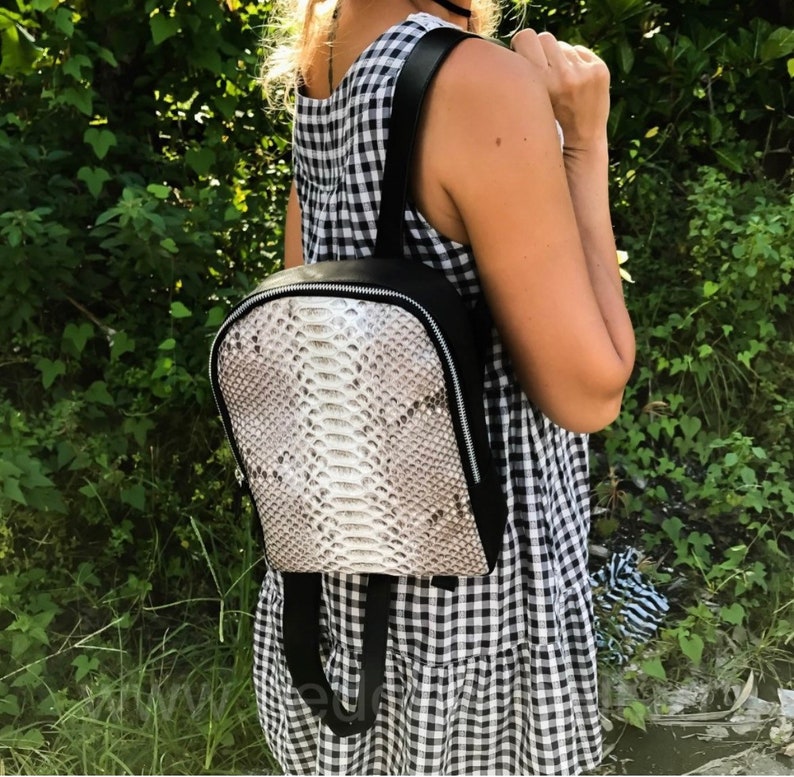 以下が含まれることがあります： 白いヘビの皮の模様がついた黒い革のバックパック。バックパックはジッパーで閉じて、調節可能なストラップが付いています。