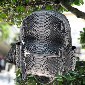 Może przedstawiać: Szary i czarny plecak ze skóry węża z kieszenią zapinaną na zamek błyskawiczny i dwoma frędzlami.