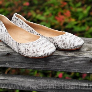 May include: A pair of white and gray patterned ballet flats. The flats have a textured, snakeskin-like appearance and a rounded toe. The soles are a light brown color. The shoes are resting on a wooden surface.