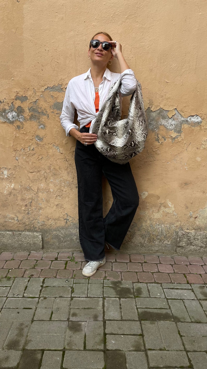 May include: A woman wearing a white button-down shirt, black pants, and white sneakers. She is holding a snakeskin handbag and wearing sunglasses.