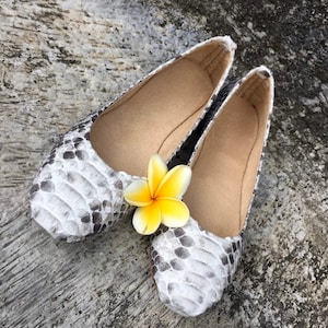May include: A pair of ballet flats with a snakeskin pattern in shades of gray and white. The shoes have a beige interior and are adorned with a yellow and white flower. The flats are displayed on a textured gray surface.