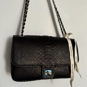 May include: Black leather handbag with a snakeskin pattern and silver chain strap. The bag features a silver clasp and a white ribbon with the text "HEDONISTBAU".