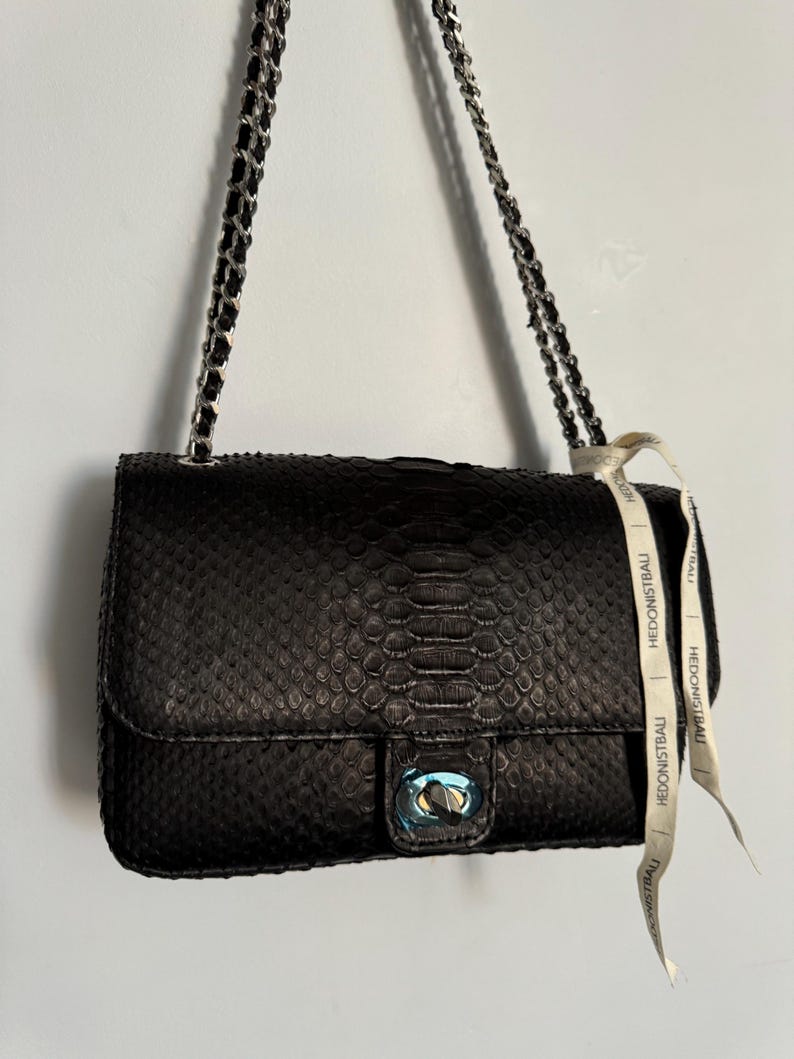 May include: Black leather handbag with a snakeskin pattern and silver chain strap. The bag features a silver clasp and a white ribbon with the text "HEDONISTBAU".