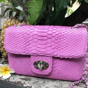 K&ouml;nnte beinhalten: Eine leuchtend pinkfarbene Schlangenlederschultertasche mit einer silbernen Kettenkette und einem silbernen Verschluss. Die Tasche hat eine strukturierte Oberfl&auml;che und einen Klappenverschluss.