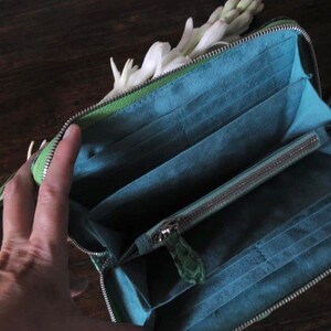 以下が含まれることがあります： ジッパーで閉じられる緑色の革製の財布が開いており、青い布地の内部には、複数のカードスロットとジッパー付きのポケットが見えます。財布は木製の表面の上に置かれています。