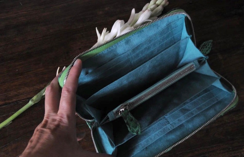 以下が含まれることがあります： ジッパーで閉じられる緑色の革製の財布が開いており、青い布地の内部には、複数のカードスロットとジッパー付きのポケットが見えます。財布は木製の表面の上に置かれています。