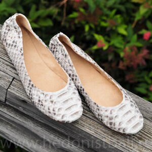May include: A pair of white and gray patterned ballet flats. The shoes have a rounded toe and a low heel. The interior of the shoe is a light tan color. The shoes are made of a textured material, possibly leather, and are displayed on a wooden surface.